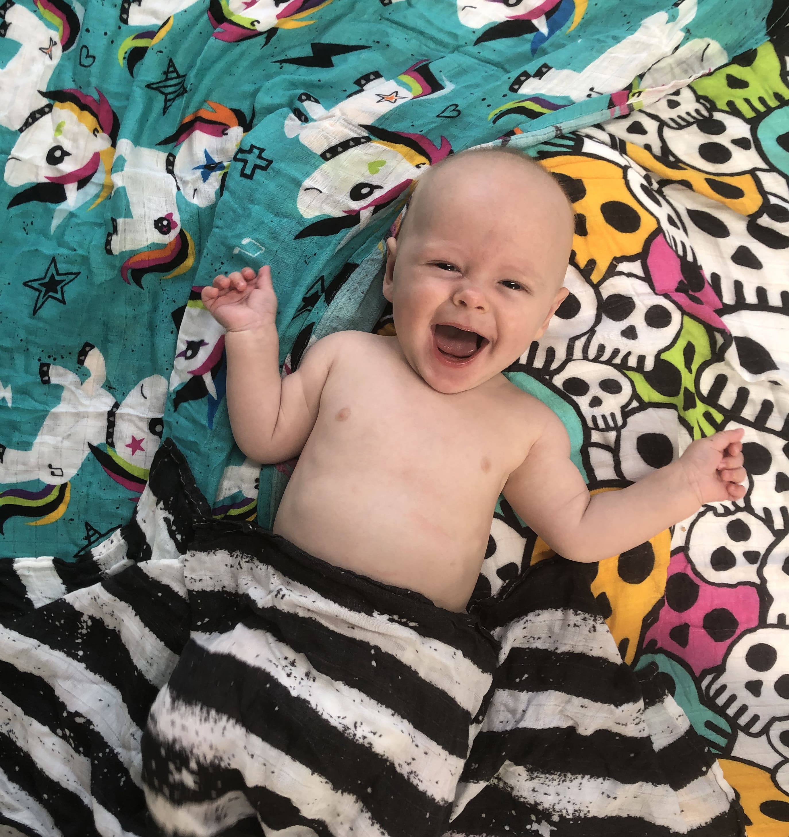 Happy baby lying on bamboo muslin swaddle blankets with unicorns and skulls